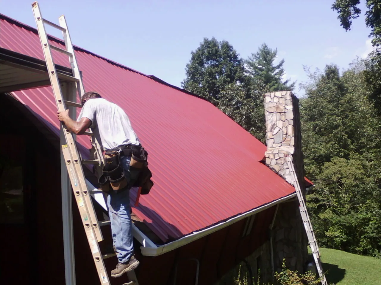 Roofer on a ladder beside a red metal roof and stone chimney — Soffit & Fascia Repair by Roofing Brothers in Slayden